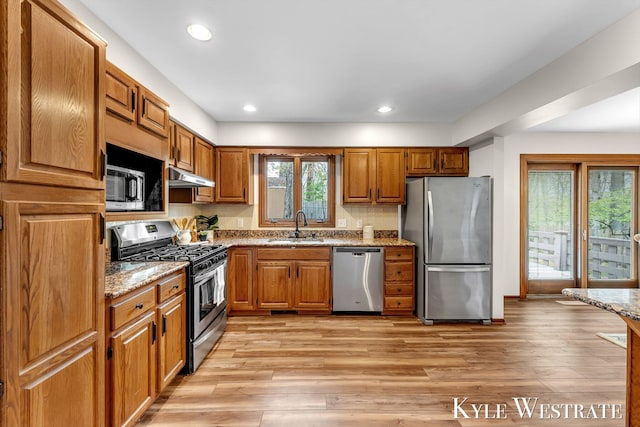kitchen with stainless steel appliances, under cabinet range hood, a sink, light wood finished floors, and brown cabinetry