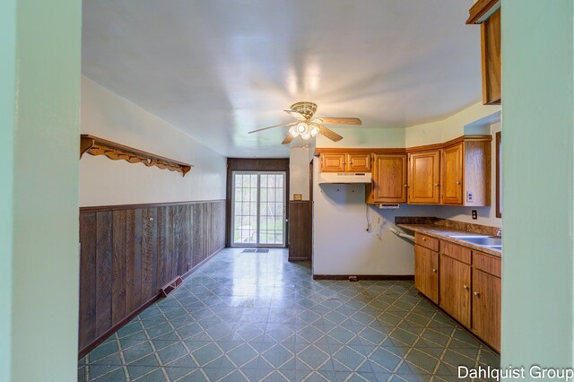 kitchen with under cabinet range hood, a sink, a ceiling fan, wainscoting, and brown cabinetry