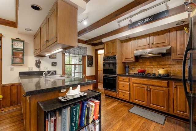 kitchen with black appliances, under cabinet range hood, a sink, wainscoting, and dark countertops