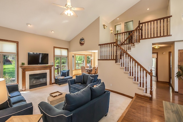 living area with high vaulted ceiling, stairs, a glass covered fireplace, a ceiling fan, and baseboards