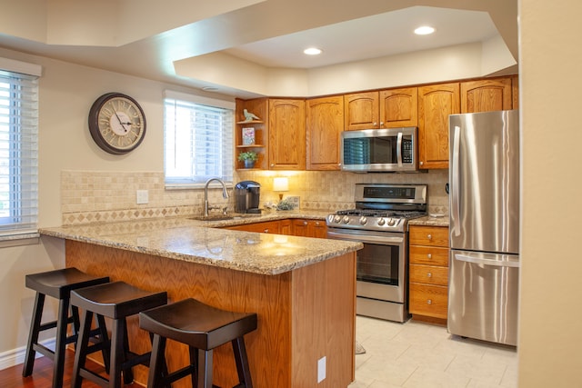kitchen with stainless steel appliances, a sink, a peninsula, light stone counters, and brown cabinetry