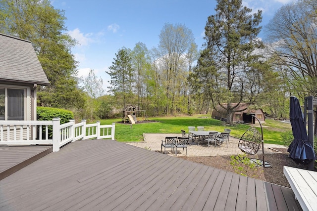 wooden terrace with a playground, a lawn, outdoor dining area, a patio, and view of wooded area