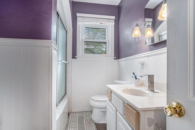 bathroom featuring a wainscoted wall, toilet, vanity, and enclosed tub / shower combo