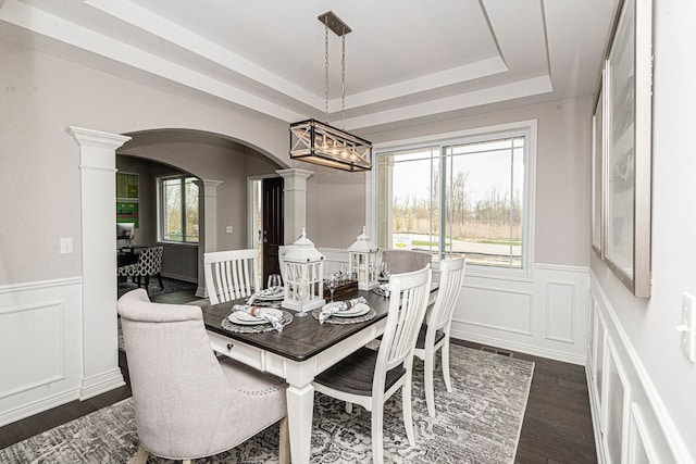 dining area featuring ornate columns, arched walkways, dark wood-style flooring, a decorative wall, and wainscoting