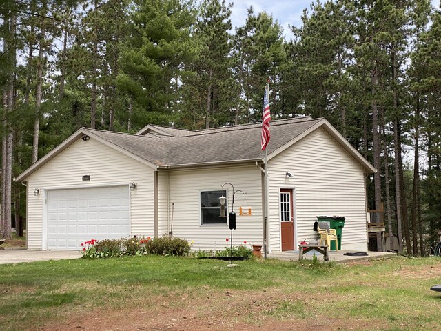 view of front of home with a garage, concrete driveway, a shingled roof, a front yard, and view of wooded area
