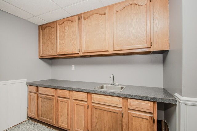 kitchen with a sink, a wainscoted wall, dark countertops, light brown cabinets, and a paneled ceiling