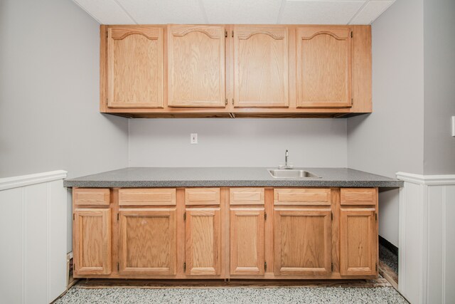 kitchen with a sink, wainscoting, dark countertops, a decorative wall, and light brown cabinets