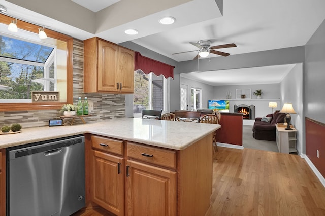 kitchen with stainless steel dishwasher, a lit fireplace, a peninsula, a ceiling fan, and decorative backsplash