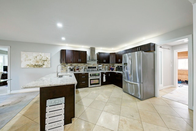 kitchen featuring appliances with stainless steel finishes, a sink, wall chimney exhaust hood, a peninsula, and light stone countertops