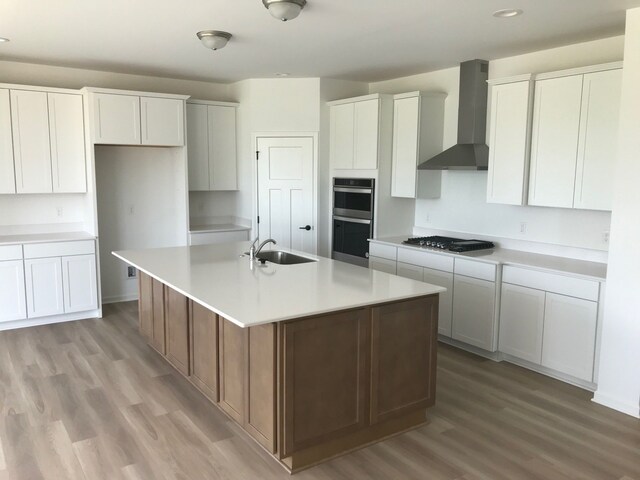 kitchen featuring wall chimney exhaust hood, double oven, a sink, gas cooktop, and a center island with sink