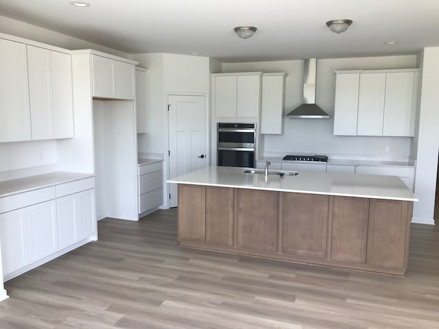 kitchen with wall chimney exhaust hood, stainless steel double oven, a sink, a kitchen island with sink, and light wood-style flooring