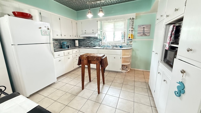 kitchen featuring freestanding refrigerator, backsplash, under cabinet range hood, light tile patterned floors, and decorative light fixtures