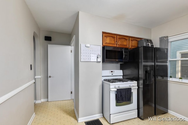 kitchen featuring white gas range, brown cabinets, arched walkways, light flooring, and baseboards