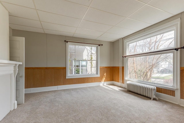 unfurnished living room featuring radiator heating unit, plenty of natural light, a drop ceiling, carpet floors, and wainscoting