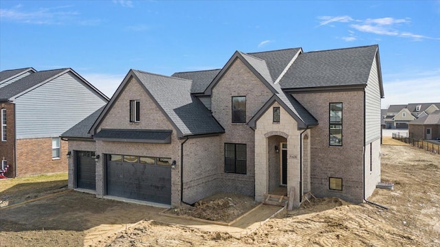 french country style house with brick siding, an attached garage, a shingled roof, and dirt driveway