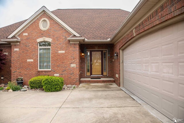 property entrance featuring brick siding, a shingled roof, driveway, and an attached garage