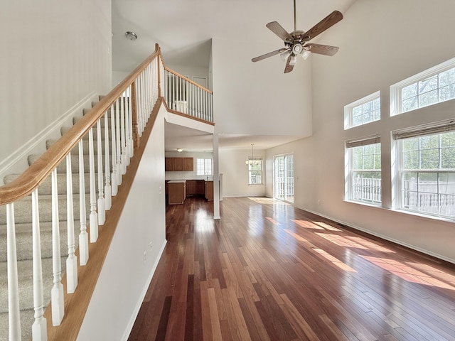 unfurnished living room featuring a towering ceiling, dark wood-style floors, plenty of natural light, stairway, and ceiling fan