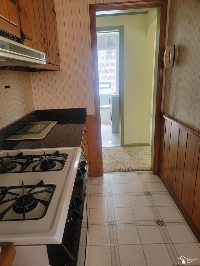 kitchen with gas stove, under cabinet range hood, wooden walls, brown cabinets, and light colored carpet