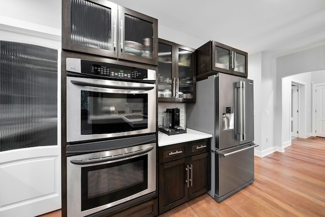 kitchen with appliances with stainless steel finishes, light wood finished floors, light countertops, glass insert cabinets, and dark brown cabinets