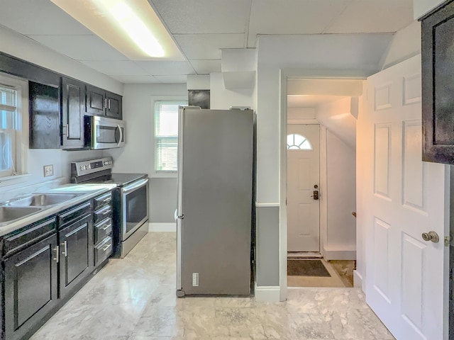 kitchen featuring stainless steel appliances, a sink, dark cabinets, a drop ceiling, and baseboards