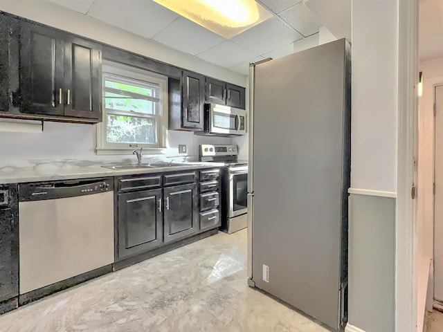 kitchen featuring stainless steel appliances, dark cabinets, a sink, a drop ceiling, and light marble finish flooring