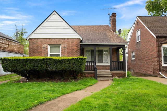 view of front of home with brick siding, a front lawn, covered porch, a chimney, and roof with shingles