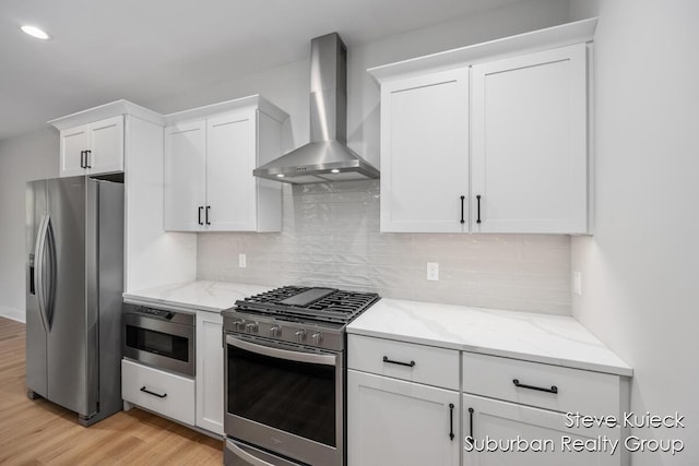 kitchen featuring appliances with stainless steel finishes, wall chimney exhaust hood, light wood finished floors, white cabinets, and light stone countertops