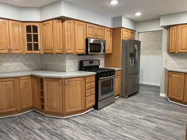 kitchen with appliances with stainless steel finishes, glass insert cabinets, wood finished floors, decorative backsplash, and recessed lighting