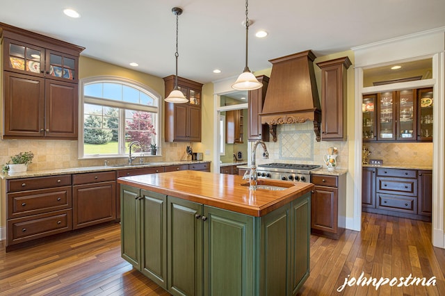 kitchen with butcher block counters, a sink, premium range hood, a kitchen island with sink, and tasteful backsplash