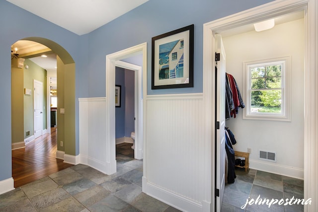 corridor with arched walkways, stone tile floors, a wainscoted wall, and baseboards
