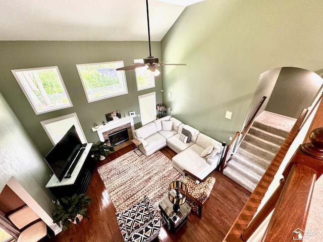 living room featuring wood finished floors, high vaulted ceiling, a fireplace with flush hearth, stairs, and ceiling fan