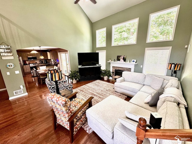 living area with high vaulted ceiling, arched walkways, dark wood finished floors, ceiling fan, and a stone fireplace