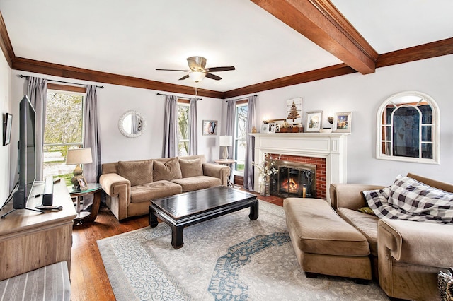 living area featuring ceiling fan, a brick fireplace, wood finished floors, ornamental molding, and beam ceiling