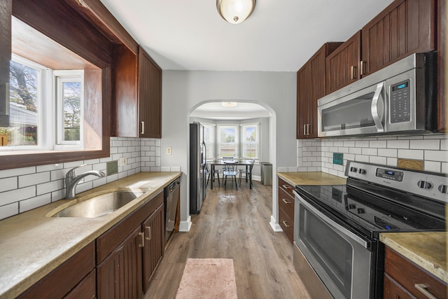 kitchen with stainless steel appliances, a sink, arched walkways, light wood-style floors, and tasteful backsplash