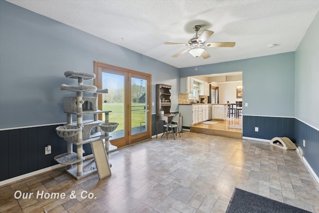 interior space with stone tile floors, a ceiling fan, a wainscoted wall, a textured ceiling, and french doors