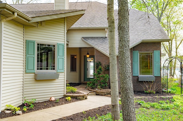 doorway to property featuring a shingled roof, a chimney, and brick siding