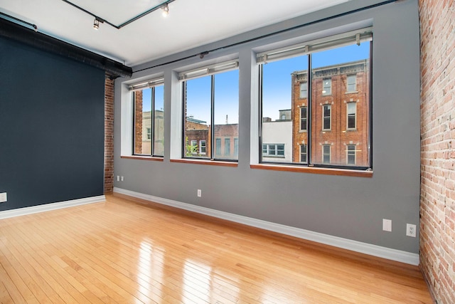 empty room with wood-type flooring, healthy amount of natural light, brick wall, baseboards, and rail lighting