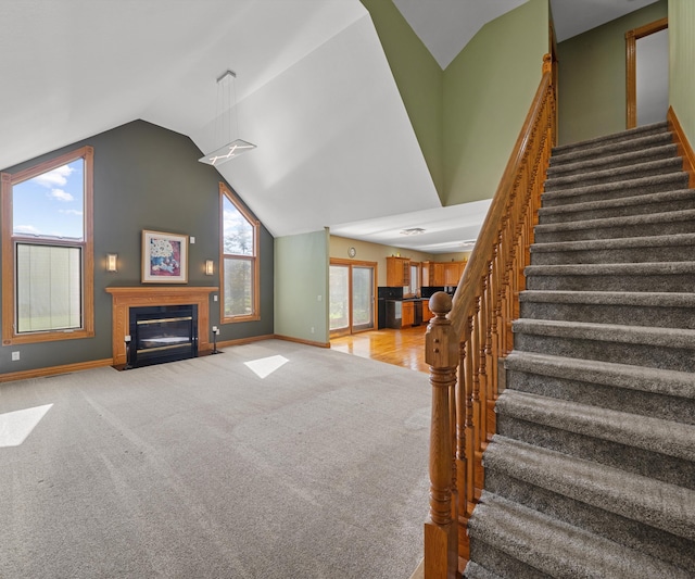 unfurnished living room featuring vaulted ceiling, stairway, a fireplace with flush hearth, light colored carpet, and baseboards