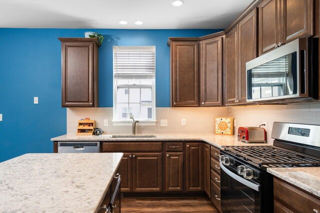 kitchen featuring stainless steel appliances, a sink, dark brown cabinets, light stone countertops, and dark wood-style floors