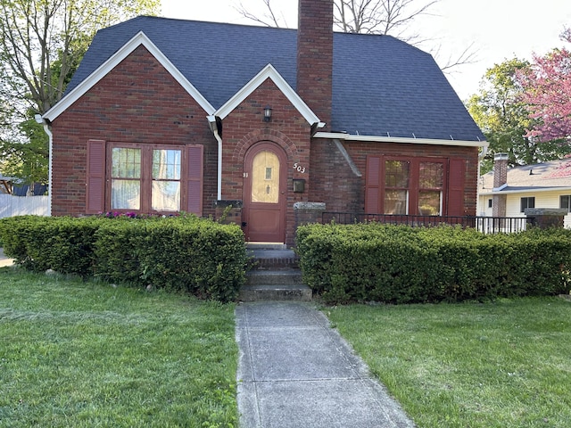 tudor-style house with brick siding, a chimney, a front lawn, and a shingled roof