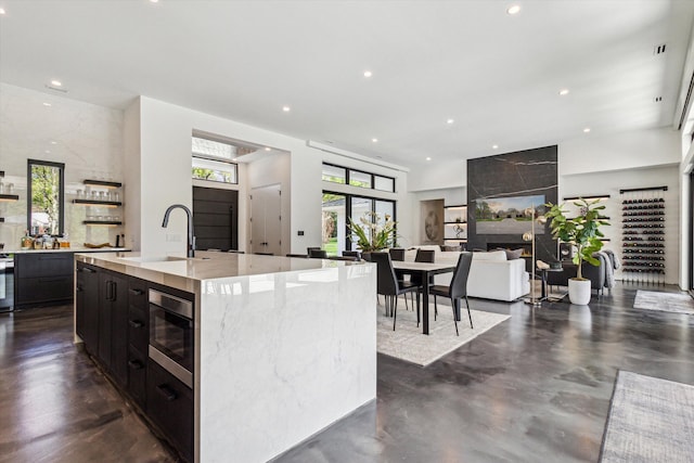 kitchen with a sink, stainless steel microwave, recessed lighting, a center island with sink, and open shelves