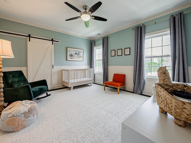carpeted bedroom with a wainscoted wall, a barn door, ceiling fan, ornamental molding, and a crib