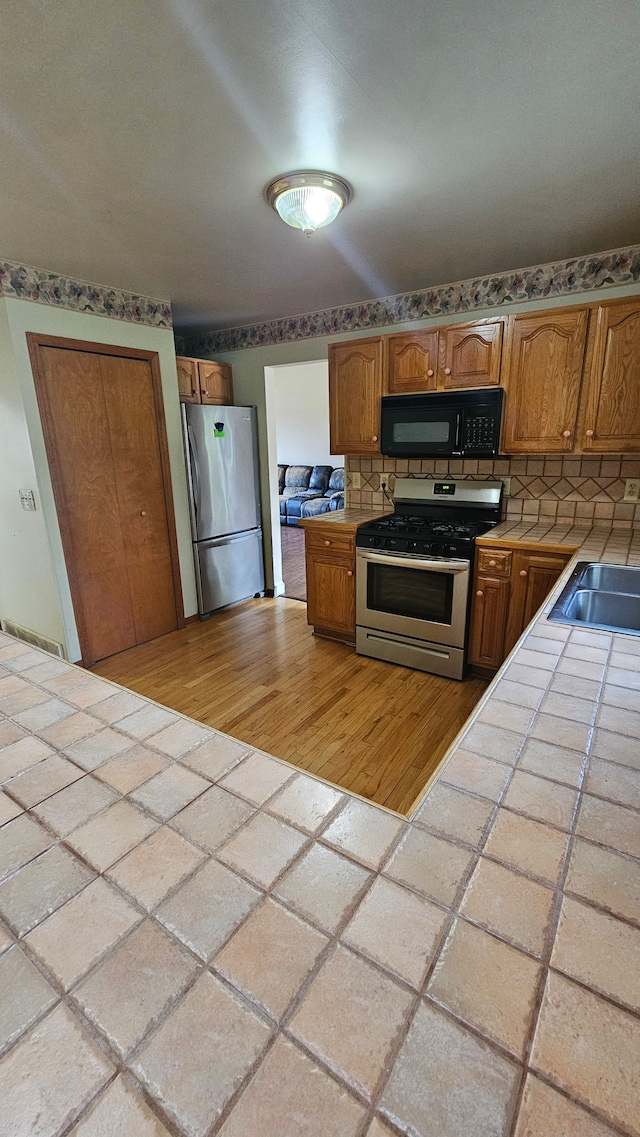 kitchen with stainless steel appliances, tile countertops, a sink, brown cabinets, and light wood-style floors