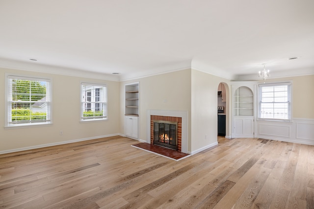 unfurnished living room featuring built in features, light wood-style floors, ornamental molding, a chandelier, and a fireplace