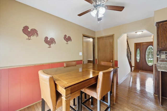 dining room with arched walkways, a ceiling fan, light wood-type flooring, and wainscoting