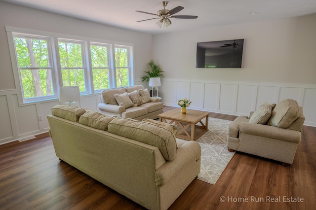 living area with a decorative wall, dark wood-style floors, a ceiling fan, and wainscoting