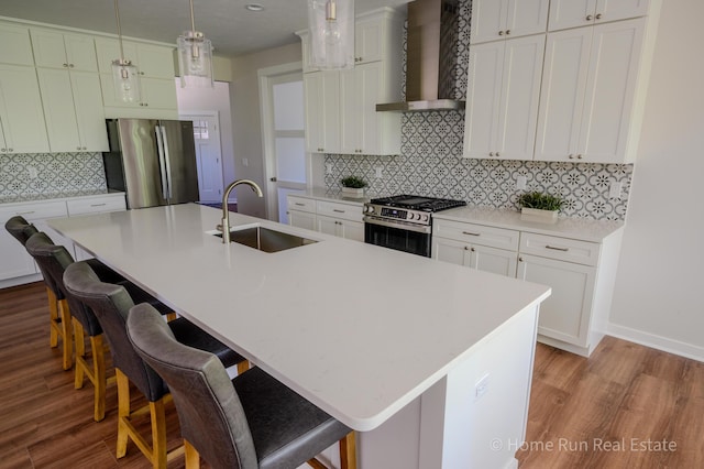 kitchen featuring wall chimney range hood, a sink, appliances with stainless steel finishes, light wood finished floors, and tasteful backsplash