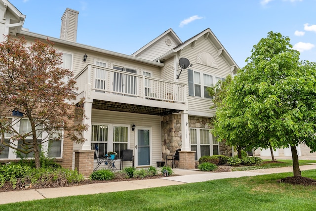 view of property featuring a balcony, stone siding, a chimney, and a front yard