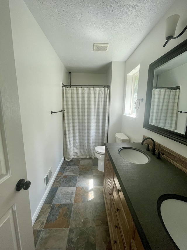 bathroom with stone finish flooring, toilet, a textured ceiling, double vanity, and baseboards