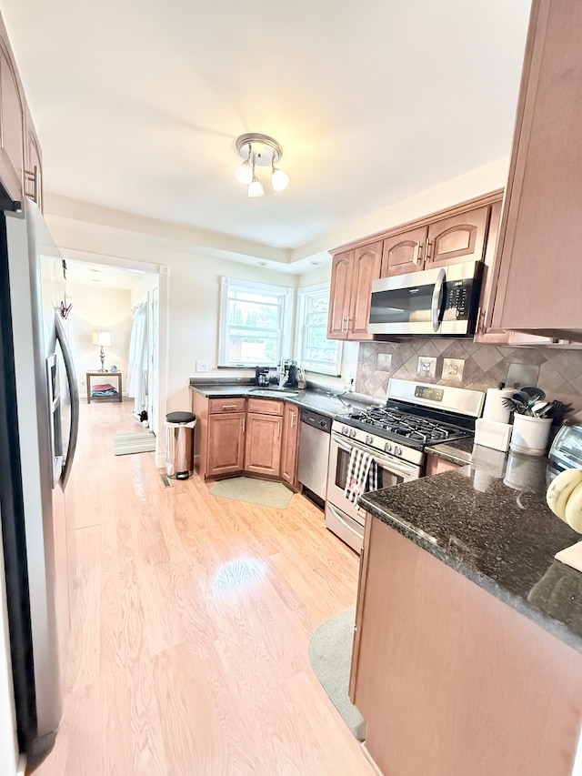 kitchen featuring stainless steel appliances, light wood-style flooring, tasteful backsplash, dark stone countertops, and a sink